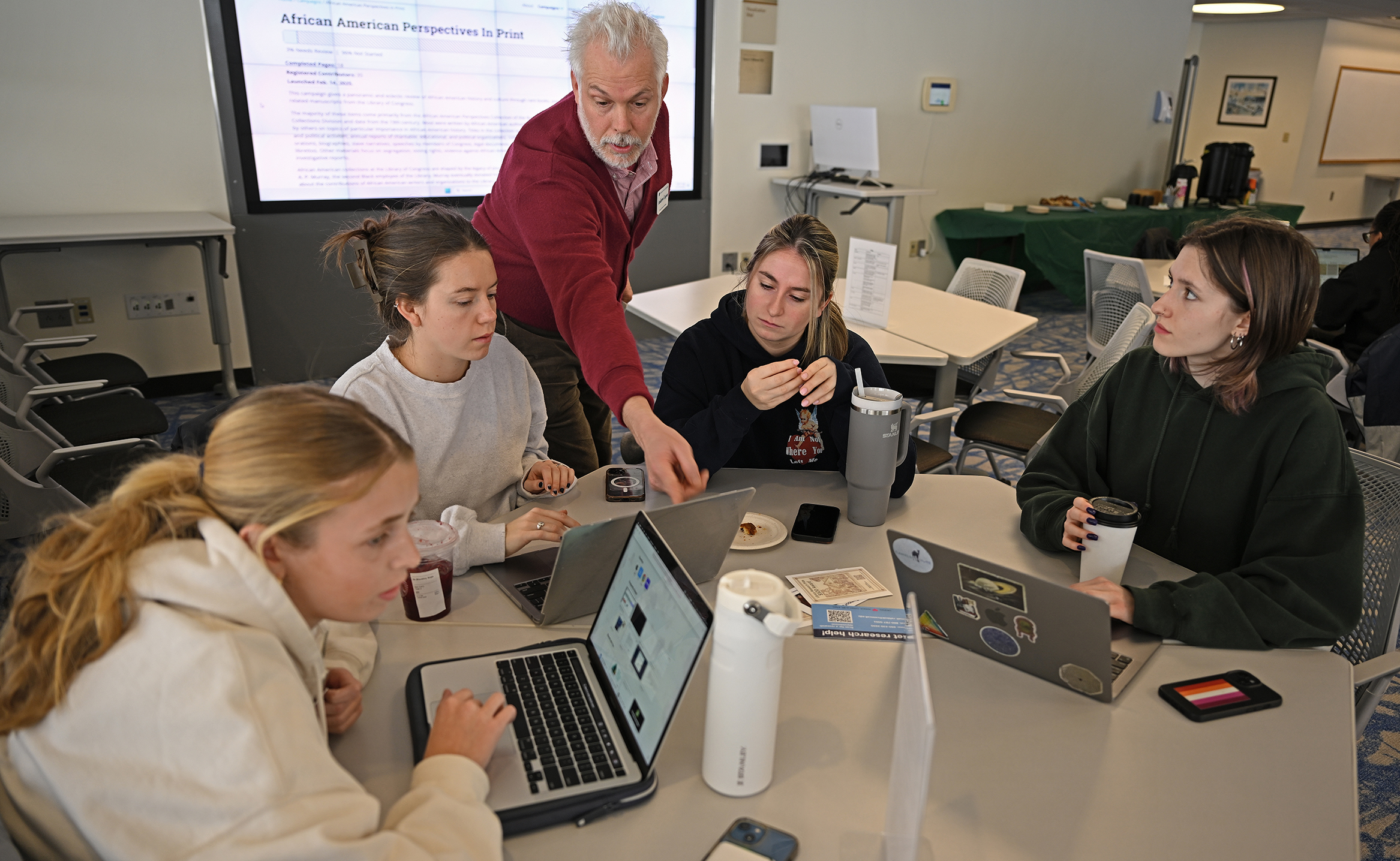 A white-haired librarian leans over a table of female students with laptop computers.