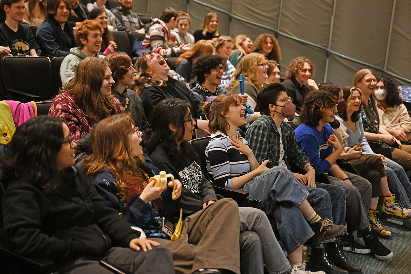 A audience of students laugh seated in an auditorium.