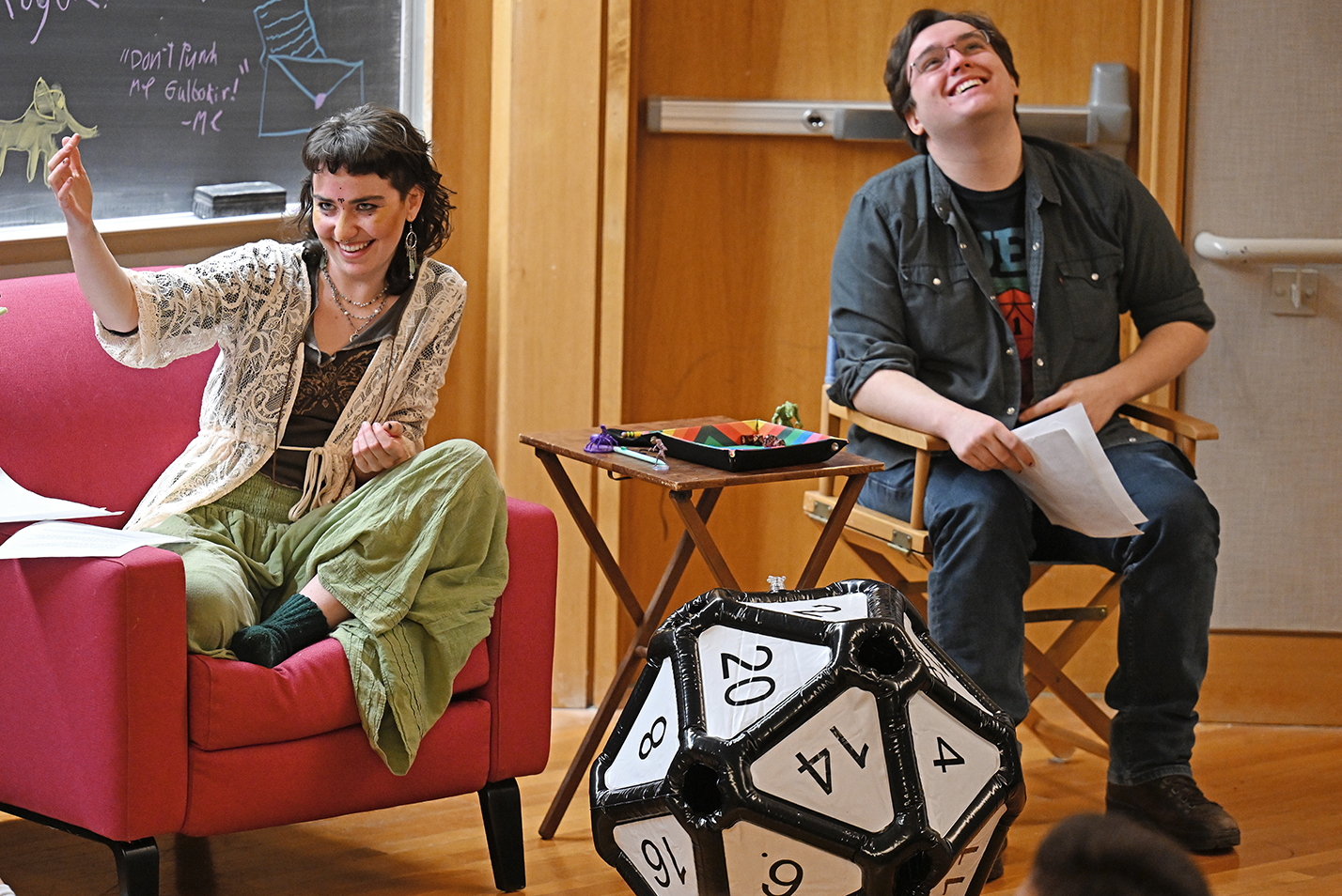 Two student dungeons and dragons players react to the game while seated next to a giant 20-sided polyhedral die.