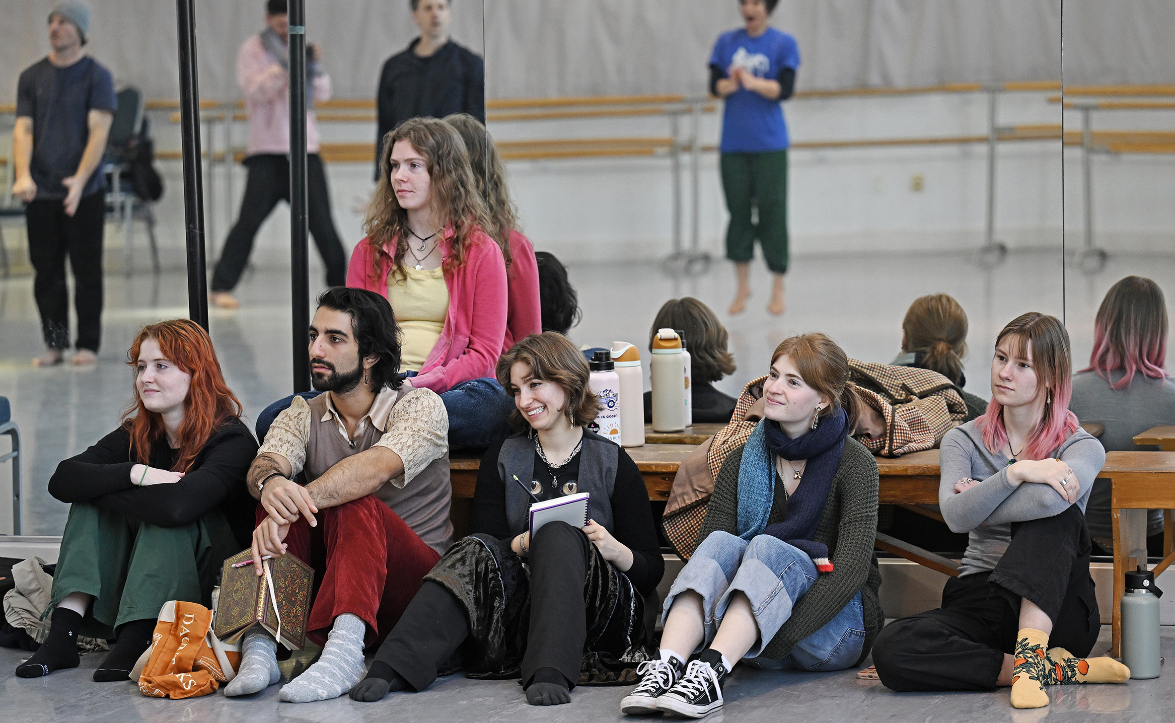 A group of dance students watch professional dancers, reflected in a mirror, rehearse a piece in a dance studio.